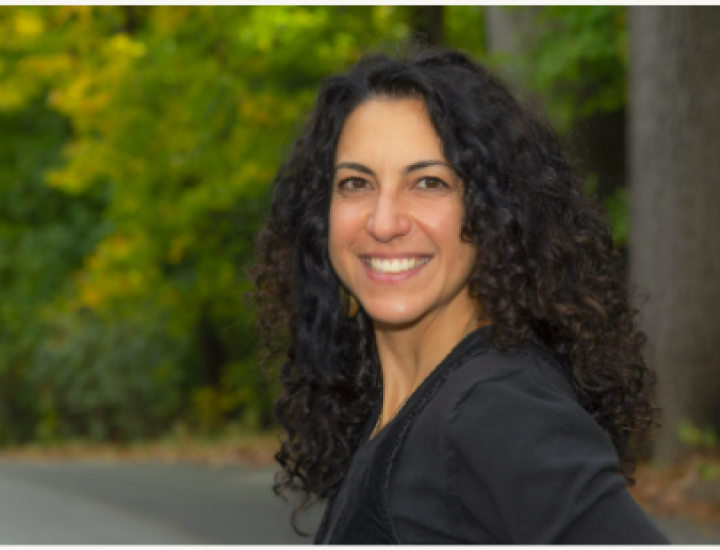 A picture of a woman with dark brown curly hair smiling at the camera, in an outdoor setting, filled with trees.