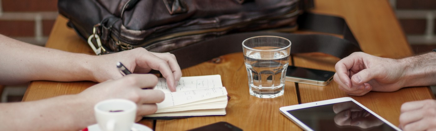 Two sets of hands, facing one another on top of a wooden table. Surrounded by personal electronics, a glass of water, espresso and briefcase. One hand is writing in a notebook.