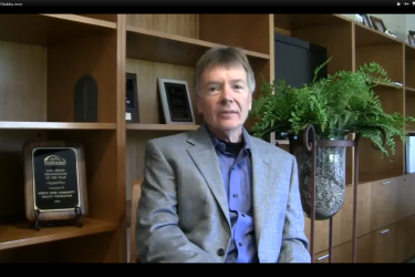 A man in a suit jacket, sitting in front of wood bookshelves and a fern.